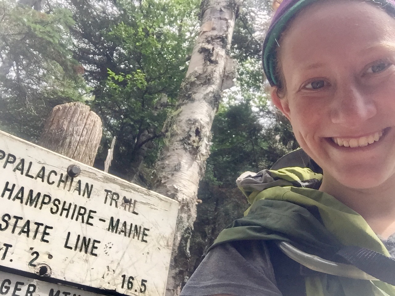 Selfie of me with a big smile in front of the small freestanding, wooden sign marking the Maine-New Hampshire state line