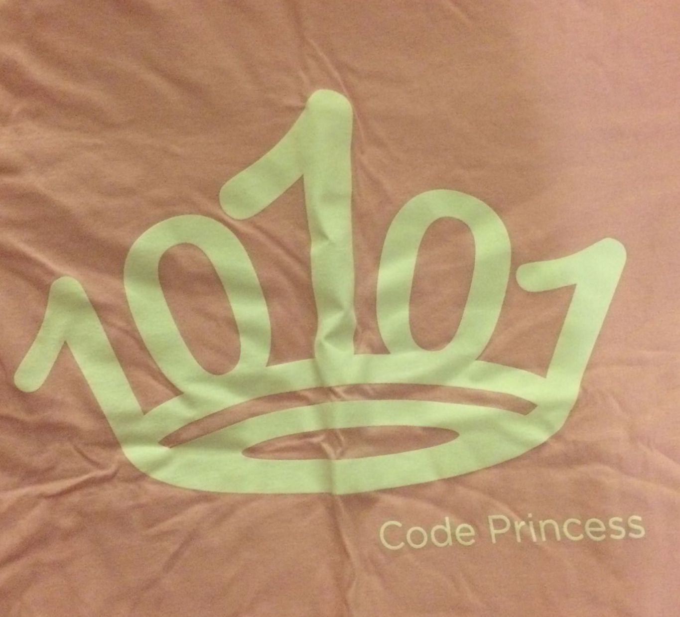 Closeup of a pink t-shirt with white lettering that says 'Code Princess' with a crown formed by 1s and 0s.