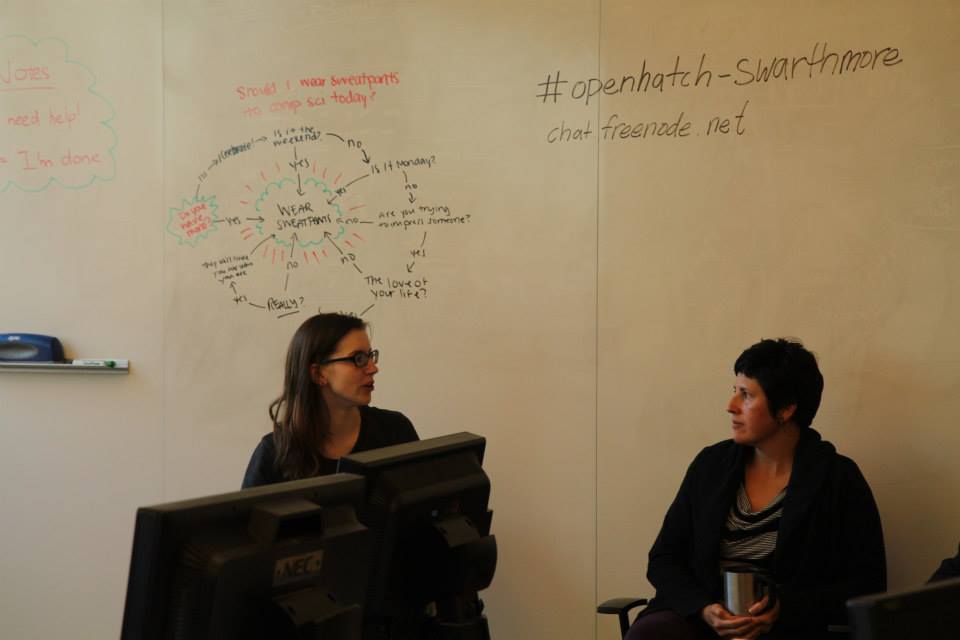 Two women facing each other with the event's Twitter tag written on the whiteboard behind them.