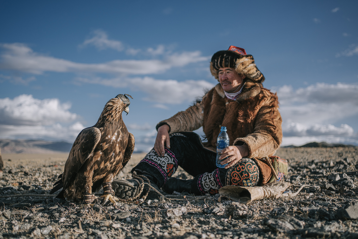 A gold eagle standing beside a traditionally dressed, sitting Kazakh Eagle Hunter in Mongolia